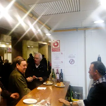 Personas comiendo chistorra y bebiendo sidra celebrando la feria de Santo Tomás dentro de una Sociedad Gastronómica.