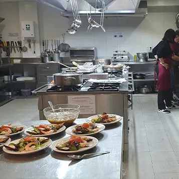 Cocina de una Sociedad Gastronómica donde dos personas están cocinando y ya hay varios platos preparados en la barra de la cocina.